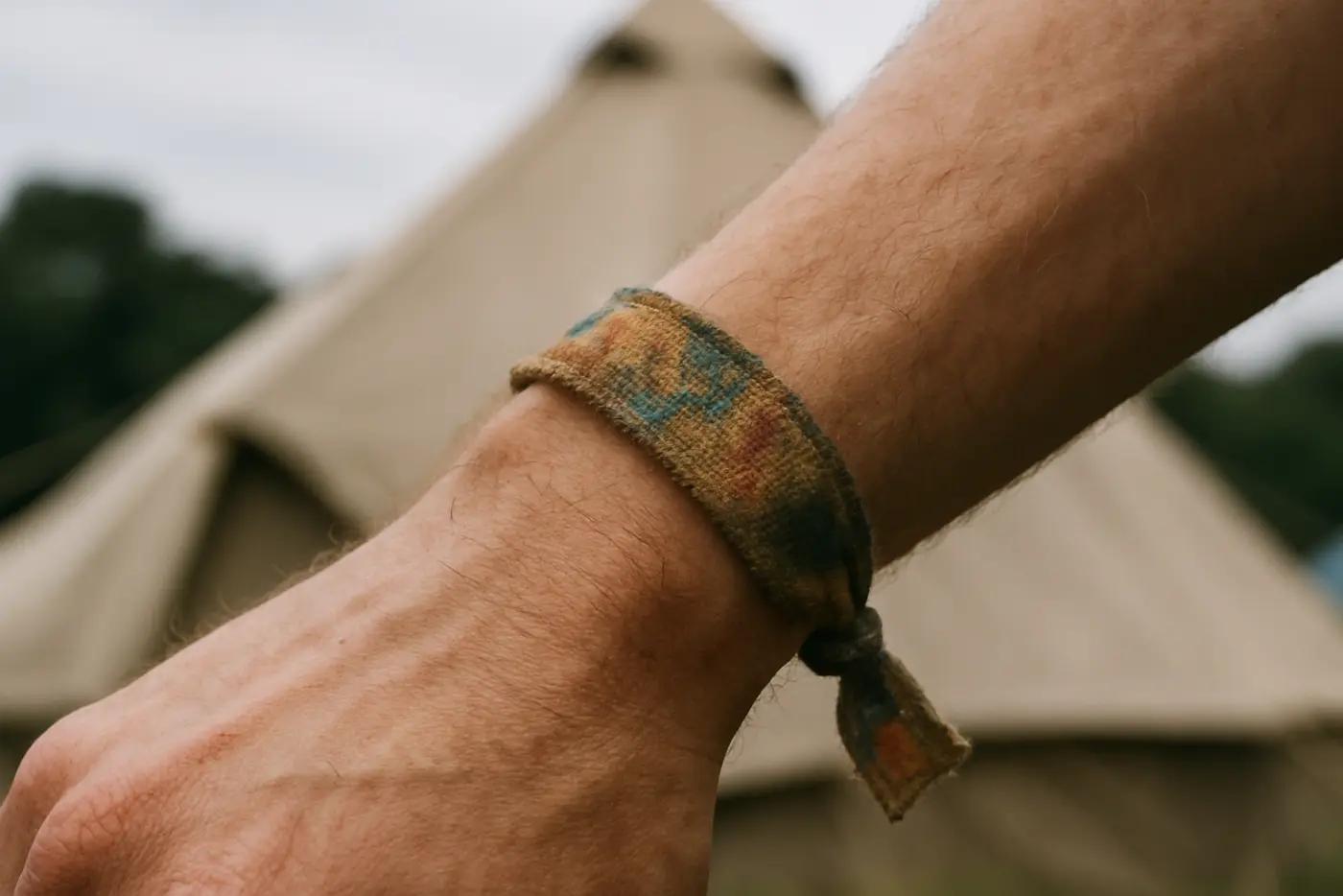 A worn woven fabric festival wristband on a wrist with tents blurred behind