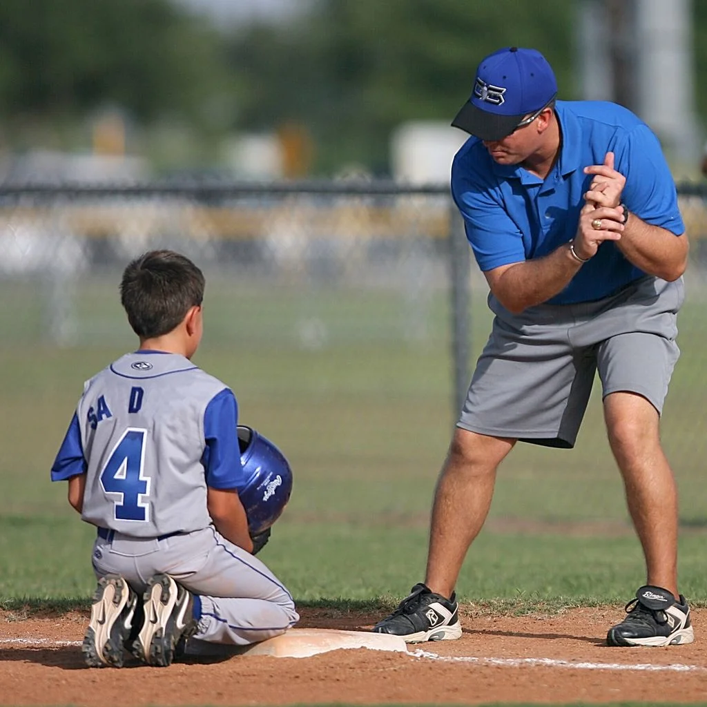 Youth baseball coach kneeling beside a young player on the field — sports team fundraising
